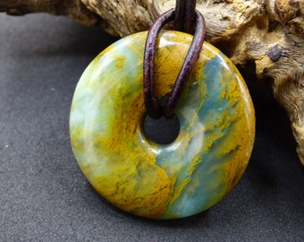 May include: A round pendant with a hole in the center, featuring a blend of yellow, blue, and green hues. The pendant is suspended from a dark brown leather cord. The stone has a marbled appearance, and the background is a dark gray surface.
