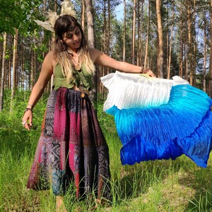 May include: A woman in a forest setting holds a large, pleated fan. The fan transitions from white to shades of blue. She wears a green crochet top and a patchwork skirt with various patterns and colors.