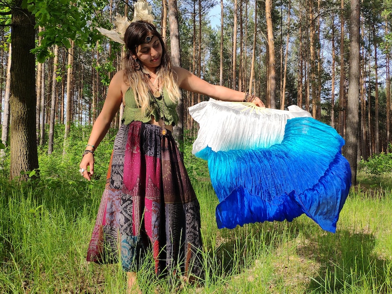 May include: A woman in a forest setting holds a large, pleated fan. The fan transitions from white to shades of blue. She wears a green crochet top and a patchwork skirt with various patterns and colors.