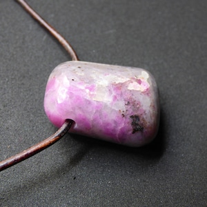 Könnte beinhalten: Ein polierter, rechteckiger Edelsteinanhänger in leuchtendem Rosa, auf einer braunen Lederschnur. Der Stein zeigt eine Mischung aus Rosa- und Weißtönen mit dunklen Sprenkeln, was eine einzigartige, natürliche Ästhetik erzeugt. Ein auffälliges Schmuckstück.