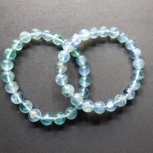 May include: Two blue fluorite beaded bracelets, each with round beads, are intertwined and laid out on a dark gray surface.
