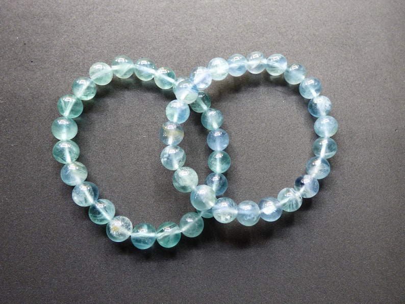May include: Two blue fluorite beaded bracelets, each with round beads, are intertwined and laid out on a dark gray surface.