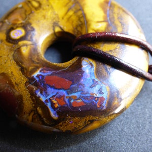 Könnte beinhalten: Ein donut-förmiger Anhänger mit einem Loch in der Mitte, gefertigt aus einem polierten Stein. Der Stein weist eine Mischung aus Farben auf, darunter Gold, Braun, Rot und Blau. Eine dunkelbraune Lederschnur ist durch das Loch des Anhängers geschlungen.