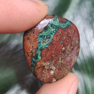 May include: A polished, heart-shaped stone with a mix of colors. The stone features a deep red-brown base with veins of vibrant teal and green. The stone is held between two fingers, showcasing its smooth, rounded form.
