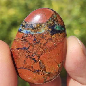 May include: A polished, oval-shaped stone featuring a complex pattern of deep red, earthy brown, and vibrant blue mineral veins. The surface is smooth and glossy, displaying a mix of jagged dark lines and patches of green and blue inclusions.