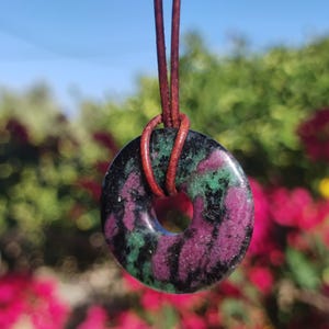 Könnte beinhalten: Halskette mit einem Anhänger in Donut-Form aus Naturstein. Die Perle hat eine Mischung aus schwarzen, grünen und violetten Farben. Der Anhänger hängt an einer braunen Lederschnur.
