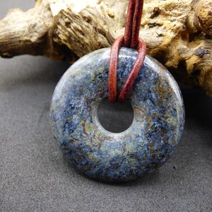 May include: A blue and yellow stone pendant with a hole in the center, strung on a brown leather cord. The stone has a marbled appearance with shades of blue, yellow, and brown. The pendant is displayed against a neutral background.