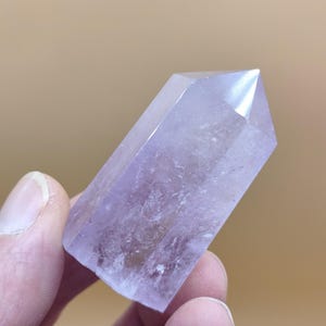 Könnte beinhalten: Ein polierter Amethyst-Kristallstab in sechseckiger Form mit einer spitzen Oberseite. Der Stein weist eine durchscheinende hellviolette Farbe mit natürlichen Einschlüssen und eine glatte, facettierte Oberfläche auf, die das Licht reflektiert.