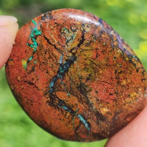May include: A polished, oval-shaped stone with a rich, earthy color palette. The surface displays a blend of reddish-brown, gold, and black hues, with streaks of turquoise and purple. The stone's texture appears to be slightly rough.