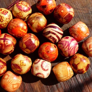Puede incluir: Una colección de cuentas de madera con intrincados patrones en tonos rojos, naranjas y beige. Las cuentas esféricas presentan varios diseños geométricos y florales. Las cuentas están dispersas sobre una superficie de madera, creando una exhibición visualmente atractiva.