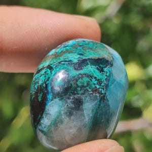 Puede incluir: Una gema pulida, de forma ovalada, con una mezcla de colores turquesa, verde azulado y negro. La piedra se sostiene entre dos dedos, mostrando su forma suave y redondeada. La superficie de la gema tiene un aspecto veteado.
