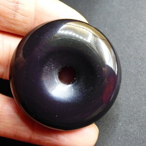 K&ouml;nnte beinhalten: Eine schwarze Obsidian-Donut-Perle mit einem Loch in der Mitte. Die Perle ist glatt und poliert.