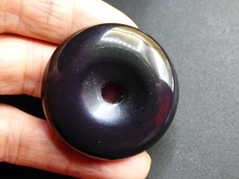 K&ouml;nnte beinhalten: Eine schwarze Obsidian-Donut-Perle mit einem Loch in der Mitte. Die Perle ist glatt und poliert.