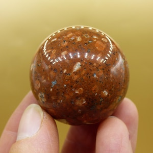 Peut inclure: Une pierre précieuse sphérique polie avec un motif tacheté de teintes brun rougeâtre, beige et bleues. L'orbe est tenue dans une main, mettant en valeur sa surface lisse et réfléchissante. Cette sphère décorative est un objet de décoration unique.