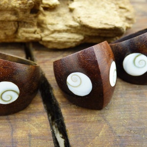 Può includere: Tre anelli in legno marrone scuro con finitura lucida. Ogni anello presenta un intarsio bianco a forma di spirale. Gli anelli sono esposti su una superficie di legno, mettendo in risalto le venature naturali e il design unico.