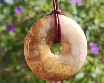 May include: A donut-shaped pendant made of beige stone, featuring a natural pattern. The pendant is suspended from a dark brown leather cord. The background is a blurred view of green foliage and purple flowers.