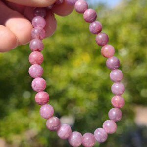 Könnte beinhalten: Ein elastisches Armband bestehend aus zahlreichen glatten, runden und durchscheinenden rosa Perlen. Die Perlen weisen subtile Unterschiede in der Farbintensität und der inneren Textur auf und sind zu einer durchgehenden kreisförmigen Schlaufe aufgereiht.