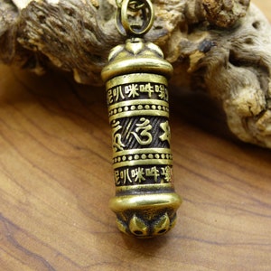 Puede incluir: Un colgante de metal dorado en forma de rueda de oración con detalles intrincados y texto grabado. El colgante está colgado de una cadena.