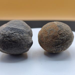 Pode incluir: Duas pedras esféricas arredondadas com superfícies rugosas e texturizadas. A pedra à esquerda é cinzento-escura com acabamento mate, enquanto a pedra à direita é de cor cinzento-acastanhada com manchas visíveis de depósitos minerais cor de ferrugem.