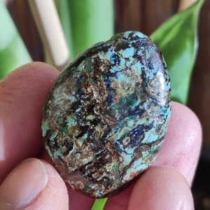 May include: A polished, oval-shaped stone with a mottled pattern of teal, brown, and dark blue. The stone has a smooth, glossy surface and is held in a hand. The natural stone is a unique decorative item.