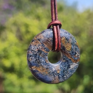 Puede incluir: Un colgante en forma de rosquilla con un patrón veteado azul, marrón y dorado, suspendido de un cordón de cuero marrón. El colgante tiene un agujero en el centro. El fondo es follaje verde borroso.