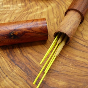 Op de afbeelding: Een houten doos met een open deksel, waaruit een bundel gele wierookstokjes tevoorschijn komt. De doos is gemaakt van donker hout met een natuurlijke houtnerf.