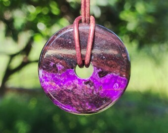 May include: A round pendant with a hole in the center, featuring a vibrant purple and brown stone. The pendant is suspended from a brown leather cord. The stone's colors and textures are visible, creating a unique accessory.
