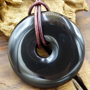 K&ouml;nnte beinhalten: Ein schwarzer Obsidian-Donut-Anh&auml;nger mit einem braunen Lederband. Der Anh&auml;nger hat eine glatte, polierte Oberfl&auml;che und ein Loch in der Mitte.