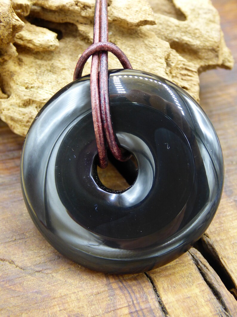 K&ouml;nnte beinhalten: Ein schwarzer Obsidian-Donut-Anh&auml;nger mit einem braunen Lederband. Der Anh&auml;nger hat eine glatte, polierte Oberfl&auml;che und ein Loch in der Mitte.