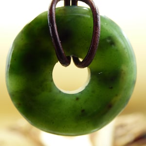 Pu&ograve; includere: Un ciondolo in giada verde con un foro al centro, appeso a un cordoncino di cuoio marrone.
