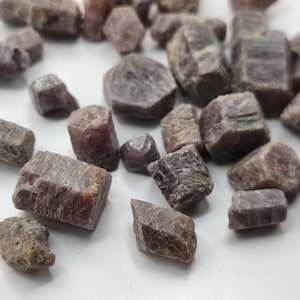 Puede incluir: Una colección de cristales de rubí en bruto y sin pulir de diversas formas y tamaños. Estas piedras preciosas naturales presentan un tono marrón rojizo terroso profundo con superficies opacas y texturizadas y estructuras cristalinas hexagonales.