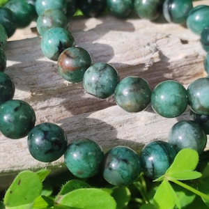 Op de afbeelding: Twee groene edelsteen armbanden met donkergroene en zwarte insluitsels. De armbanden zijn gemaakt van ronde kralen en zijn op elkaar gestapeld. De armbanden liggen op een stuk drijfhout met groene klaverblaadjes op de voorgrond.