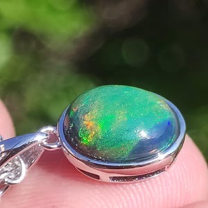 May include: A close-up of a pendant featuring a vibrant, oval-shaped opal. The opal displays a mix of green, blue, and orange colors within a silver setting. The pendant is attached to a silver chain, showcasing the gemstone's iridescent qualities.