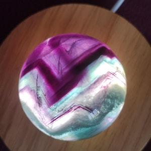 Könnte beinhalten: Eine violette und grüne Fluoritkugel auf einem Holzsockel. Die Kugel wird von innen beleuchtet, wodurch die komplizierten Muster des Kristalls hervorgehoben werden.