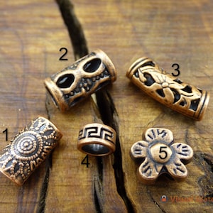 Puede incluir: Cinco cuentas de metal de color cobre con diferentes diseños. Las cuentas están sobre una superficie de madera. Las cuentas están numeradas del 1 al 5.