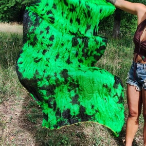 May include: A vibrant green and black tie-dye silk fan. The fan is held open, showcasing the flowing fabric's pattern. The edges are trimmed with a gold border. The fan is being held outdoors in a grassy area.