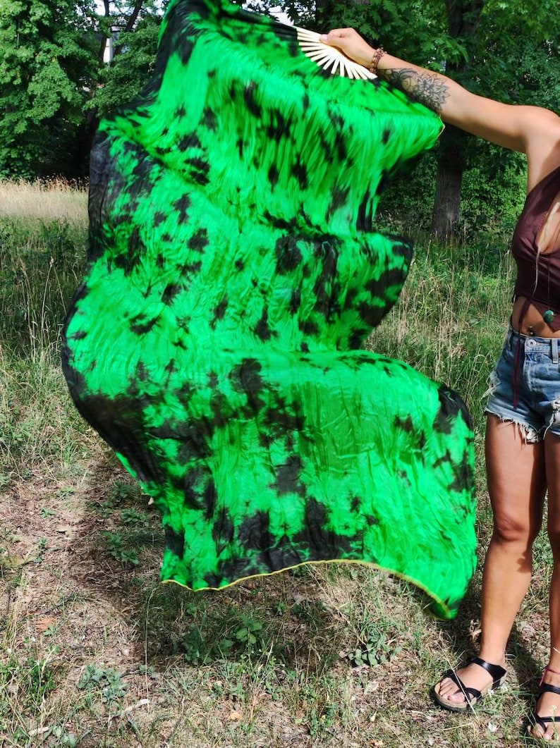 May include: A vibrant green and black tie-dye silk fan. The fan is held open, showcasing the flowing fabric's pattern. The edges are trimmed with a gold border. The fan is being held outdoors in a grassy area.