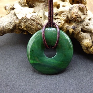 Pu&ograve; includere: Un ciondolo a forma di ciambella in giada verde con un cordoncino in pelle marrone. Il ciondolo ha una superficie liscia e lucida e un foro al centro.