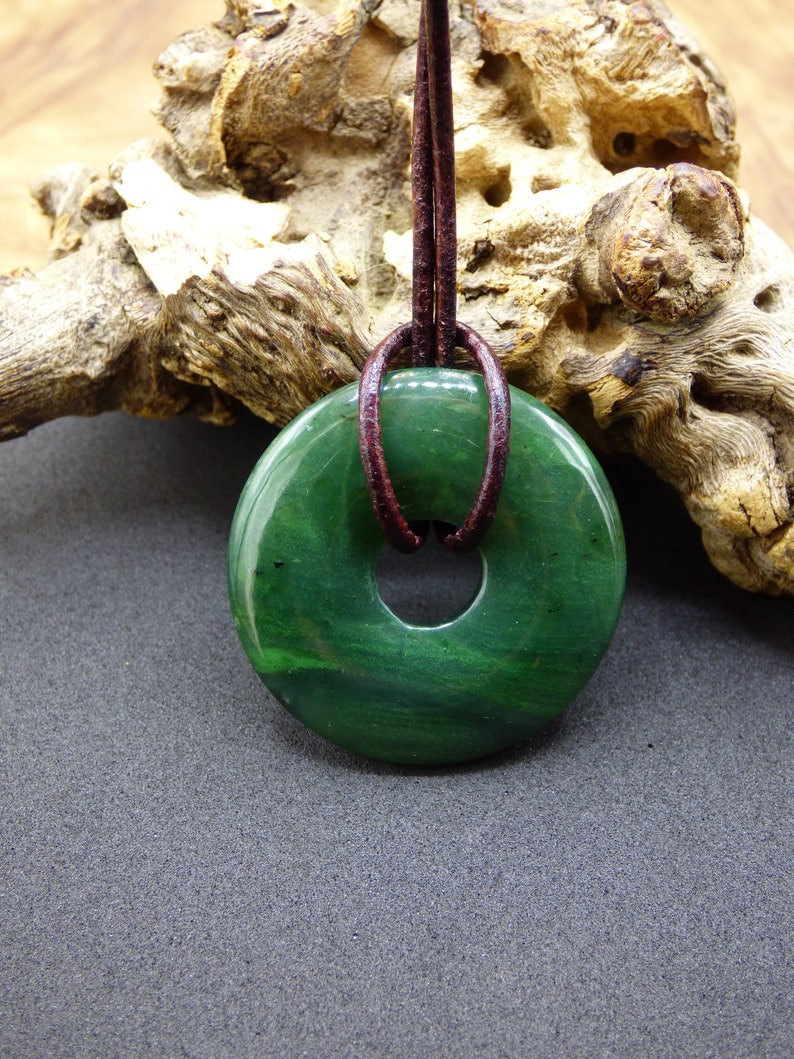 Pu&ograve; includere: Un ciondolo a forma di ciambella in giada verde con un cordoncino in pelle marrone. Il ciondolo ha una superficie liscia e lucida e un foro al centro.