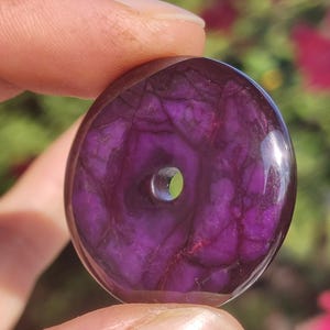 May include: A round, polished purple stone with a central hole. The stone's surface displays intricate veining patterns in darker shades of purple. The object is held between fingers, with a blurred background of green and pink.