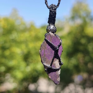 May include: A pendant featuring an irregular, flat piece of purple stone with brown veining, secured by black cord macrame wrapping. A silver-toned metal bead with an intricate cutout pattern sits above the stone, attached to a twisted brown cord.