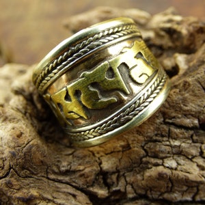 Puede incluir: Un anillo ancho de color latón con detalles intrincados. El anillo presenta una banda de diseño trenzado a lo largo de los bordes superior e inferior, con un panel central que muestra caracteres grabados. El anillo está sobre una superficie de madera texturizada.