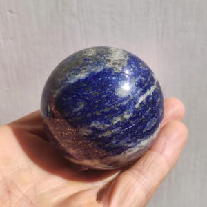 Op de afbeelding: Een gepolijste blauwe en witte lapis lazuli bol met gouden vlekken. De bol is glad en heeft een hoogglans afwerking.