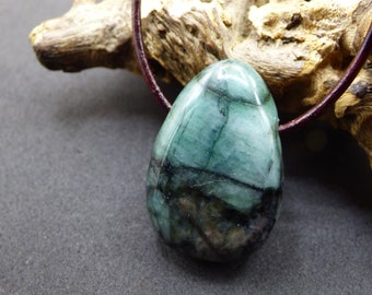 May include: A polished, teardrop-shaped emerald pendant featuring deep green and black inclusions, suspended from a dark brown leather cord. The stone has a smooth, glossy surface with visible natural veining and dark mineral patterns throughout.