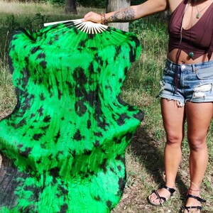May include: A large, open fan with a vibrant green and black tie-dye pattern. The fan is held open, revealing its full size and design. The fan's handle is made of light-colored wood.