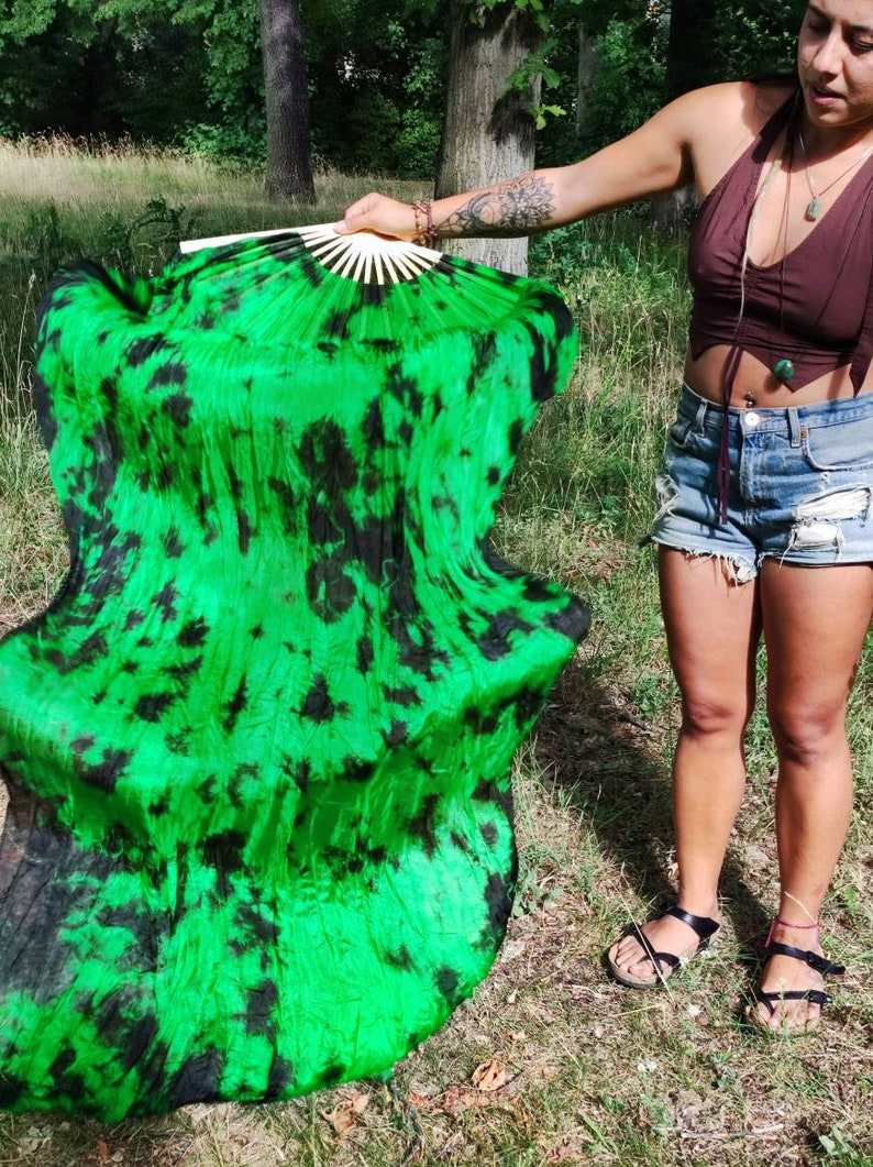 May include: A large, open fan with a vibrant green and black tie-dye pattern. The fan is held open, revealing its full size and design. The fan's handle is made of light-colored wood.