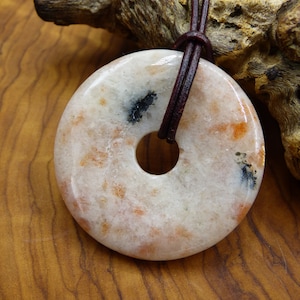 Può includere: Un ciondolo circolare in pietra del sole con una superficie lucida e liscia, caratterizzato da tonalità screziate color crema, pesca e arancio con piccole inclusioni scure. La pietra ha un foro centrale ed è appesa a un cordino in pelle.