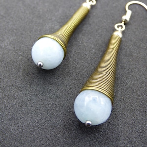 May include: A pair of earrings featuring a light blue, spherical bead at the bottom, topped with a brass-colored, cone-shaped wire wrap. The earrings have silver-colored ear wires. The earrings are displayed on a dark gray surface.