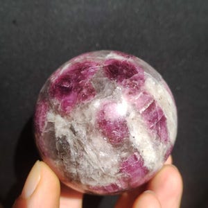 Könnte beinhalten: Eine polierte Kugel aus rosa und weißem Turmalin. Der Edelstein weist eine Mischung aus undurchsichtigen weißen und durchscheinenden rosa Abschnitten auf, wodurch ein marmorierter Effekt entsteht. Die Kugel wird in einer Hand vor dunklem Hintergrund gehalten.