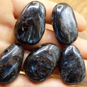 May include: Six polished, dark blue-black gemstones with iridescent blue and silver streaks. The stones are smooth and oval-shaped, with one stone having a drilled hole. The gemstones are held in a hand.